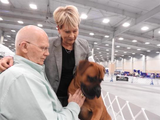 An elderly man shares a moment with a dog, aided by a woman at a dog show.