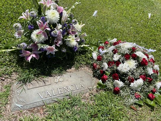 Colorful floral arrangements are laid on a gravestone in a peaceful cemetery setting.