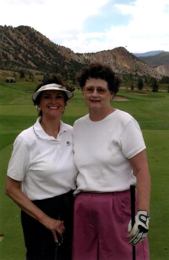 Two women are smiling and posing on a golf course surrounded by mountains on a sunny day.