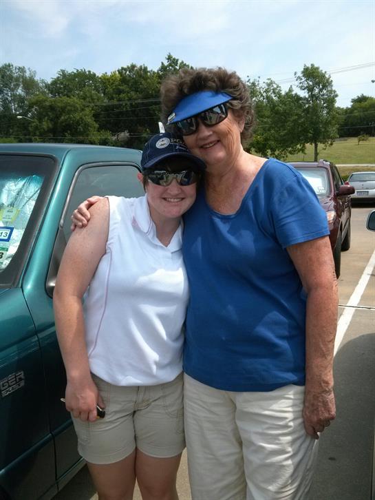 Two women smile and embrace near a parked vehicle under a clear blue sky, enjoying a sunny day.