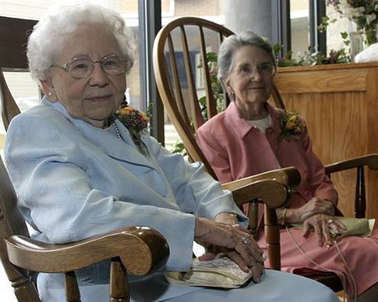 Two elderly women relax in rocking chairs, sharing a serene moment in a warm indoor space.