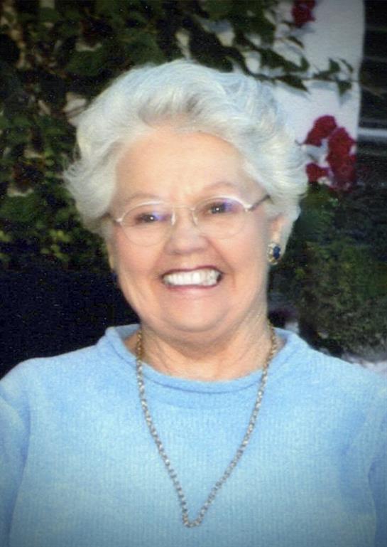 An elderly woman smiles joyfully against a colorful floral backdrop in bright sunlight.