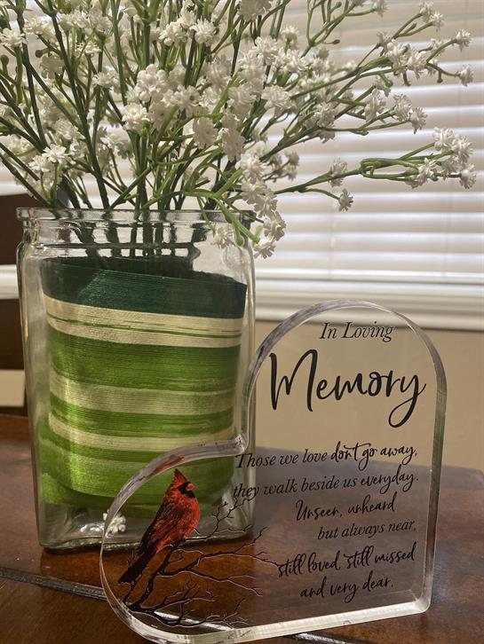 A glass vase filled with white flowers sits beside a memory plaque on a wooden table.