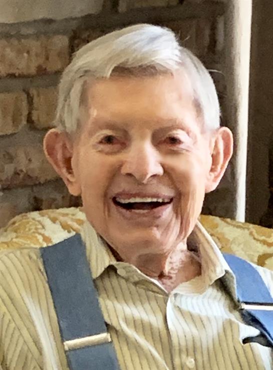A senior man with gray hair and suspenders smiles joyfully while seated in a cozy chair.