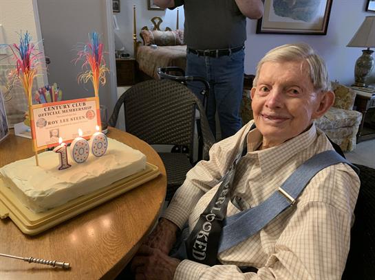 An elderly man smiles next to a birthday cake with candles and decorations.