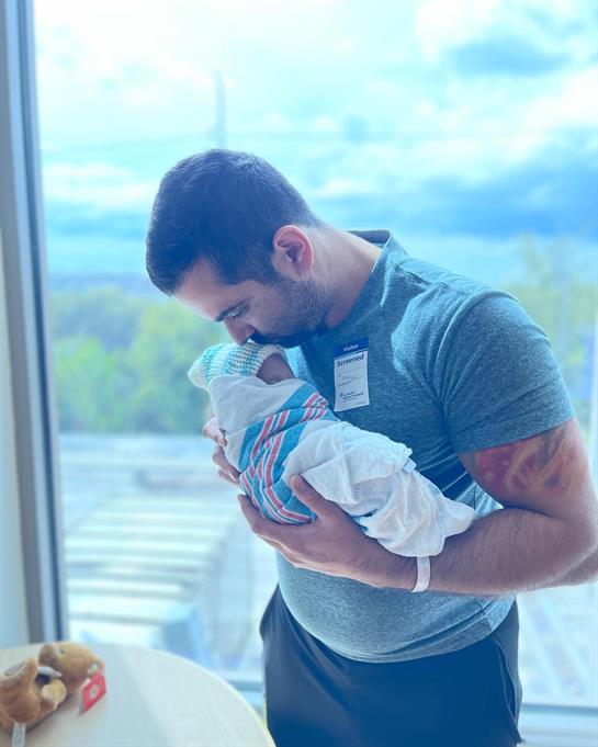 A father holds his newborn in a hospital setting, sharing a tender moment by the window.