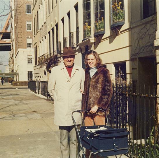 A couple stands smiling on a city street, surrounded by beautiful buildings and vibrant plants.