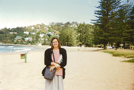A woman in casual attire smiles while standing on a beach with lush greenery in the background.