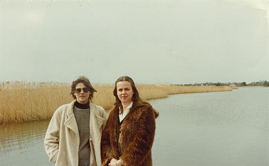 Two women in warm clothes stand by a calm body of water on a chilly autumn day.