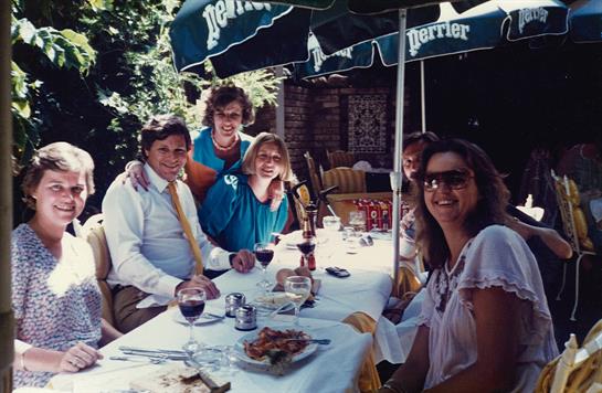 Group of friends laughing and sharing food and drinks at a shaded table during a warm day.
