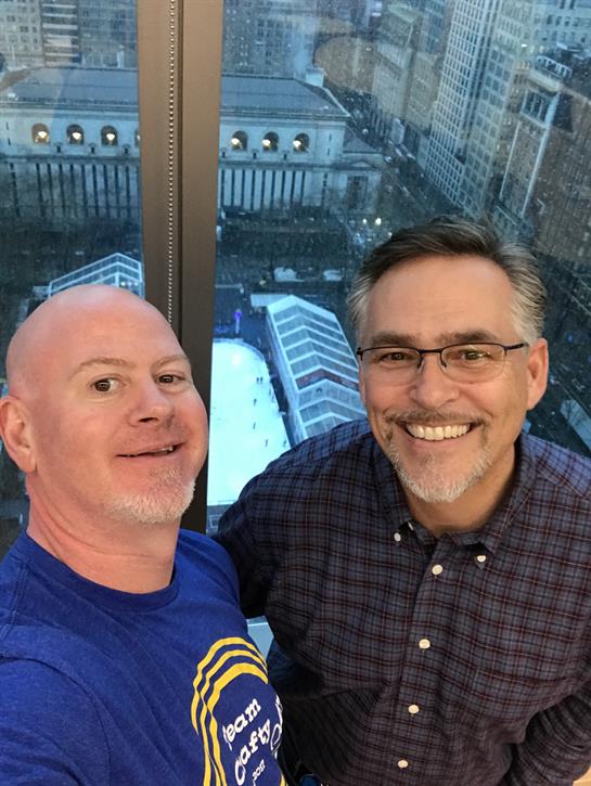 Two men smile for a cheerful selfie while enjoying an incredible city view from a tall building.