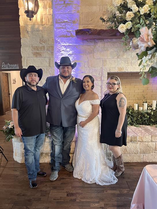 A couple in formal attire poses proudly with friends in a floral-decorated venue.