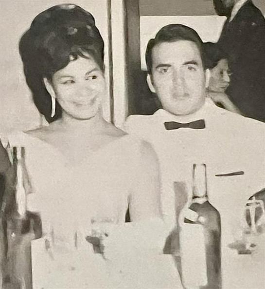 A couple smiles and poses together at a lavish dinner party surrounded by elegant decor.