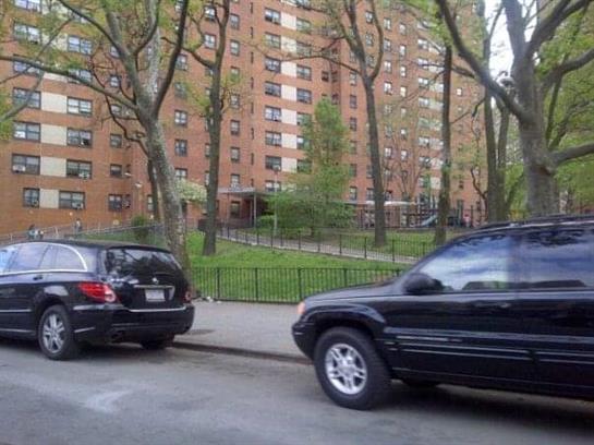Parked cars line the street alongside tall residential buildings amid trees and greenery.