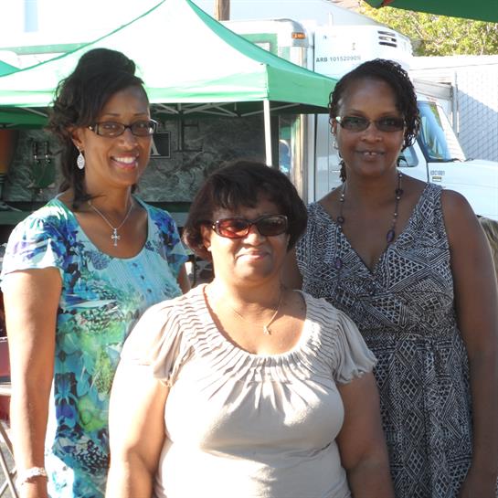 Three women pose together, smiling under green canopies at an outdoor market.