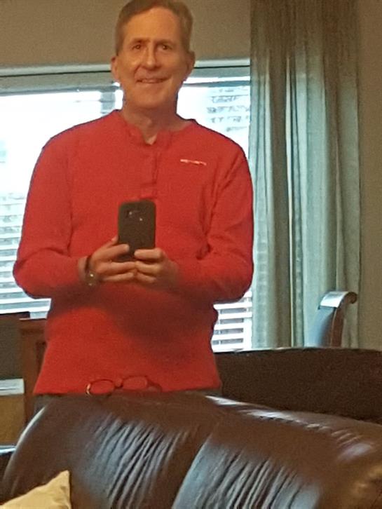 Individual stands in a well-lit room, smiling while capturing a picture with their phone.