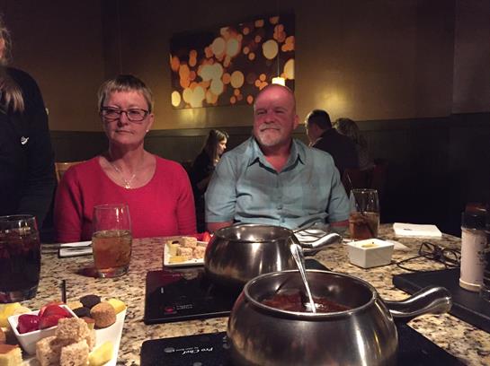 Two people sit at a table with fondue pots, surrounded by friends at a warm restaurant.