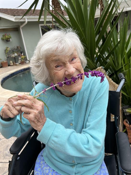 Senior woman with white hair playfully holds a flower near her mouth, smiling happily.