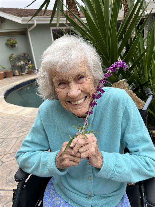 An elderly woman in a wheelchair smiles brightly while holding a purple flower by a poolside.