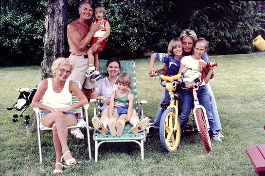 A family enjoys a sunny day in the garden, with children playing and adults lounging in chairs.