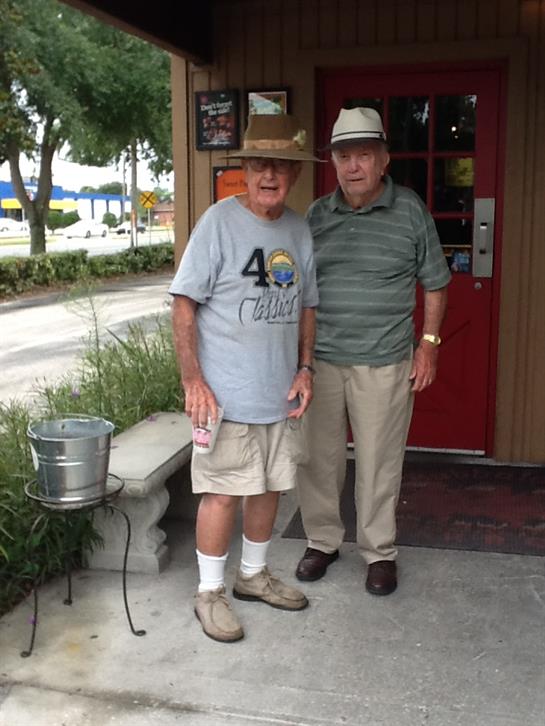 Two men stand side by side, smiling outside a restaurant with casual attire under bright sunlight.