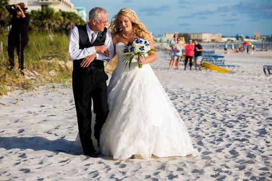 A bride in a white gown walks with her father on a sandy beach, celebrating their special day.
