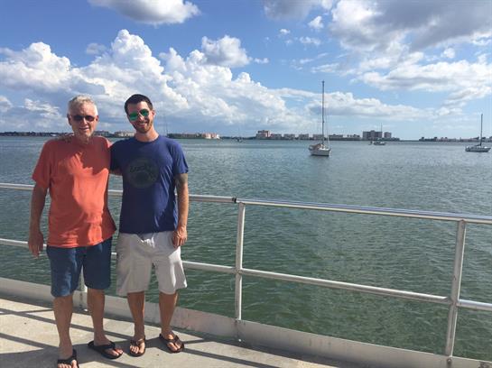 Two men smile on a pier under a bright sky, with a sailboat nearby.