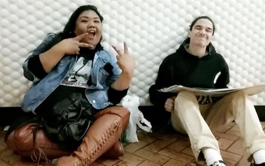 Two friends sit together, one posing with a playful gesture, while the other sketches nearby.