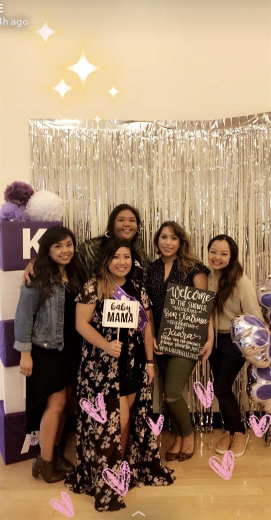A group of friends gathers for a baby shower, smiling and holding signs, surrounded by decorations.