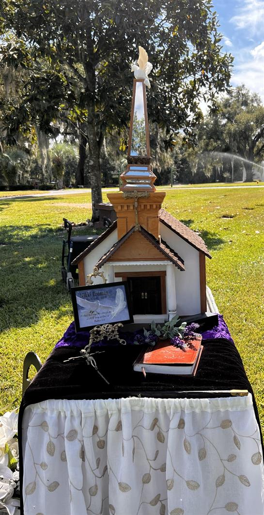 A detailed miniature church is showcased on a table surrounded by greenery in bright sunlight.