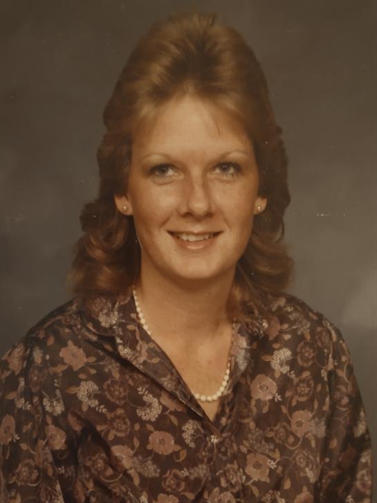 A young woman with curly hair is smiling while wearing a floral blouse in a studio.