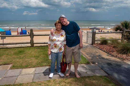A happy couple stands together by the beach, smiling and enjoying time under a sunny sky with waves.