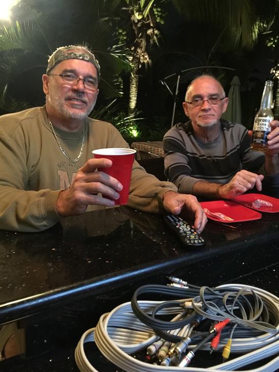 Two men sit at a bar, smiling and enjoying beverages in a warm, outdoor atmosphere at night.