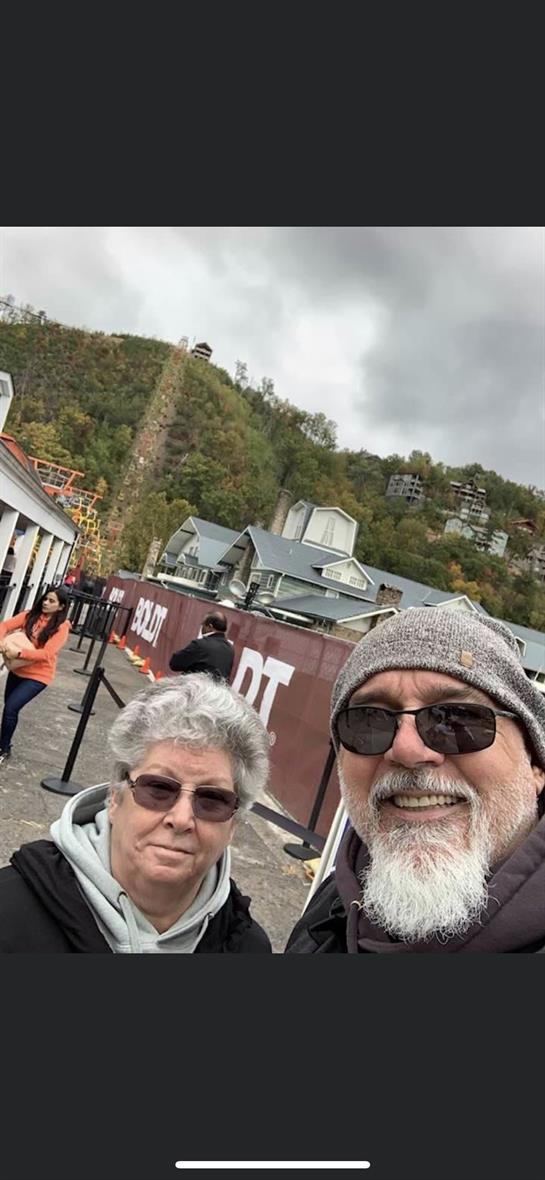 A couple poses for a selfie at a mountain resort while others socialize nearby on a cloudy day.