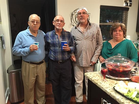 Four senior friends stand together smiling and enjoying drinks in a warm kitchen setting.