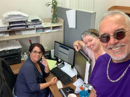 Three colleagues interact at their desks, balancing work tasks with friendly banter and smiles.