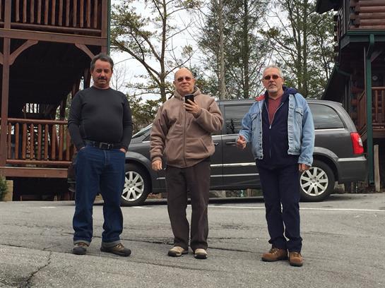 Three men pose together outdoors near a parked car, enjoying a moment in nature.