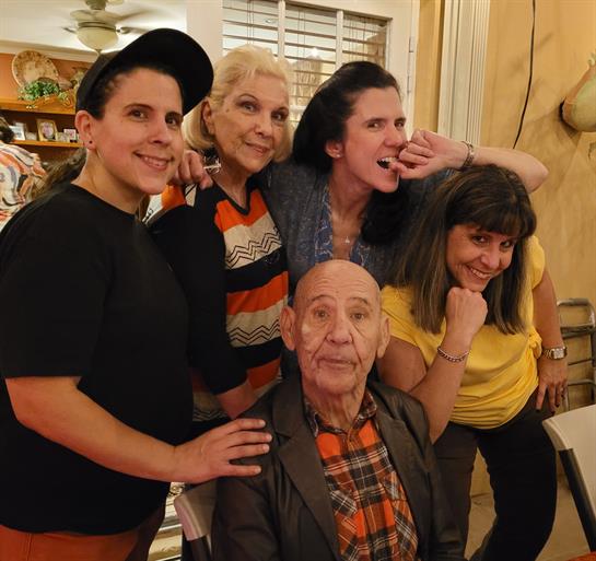 A group of five friends poses joyfully with an elderly man in a warm indoor setting.