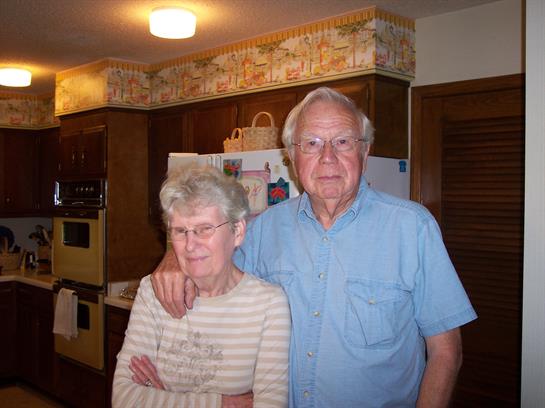 An elderly couple stands together in their home kitchen, showing affection and togetherness.