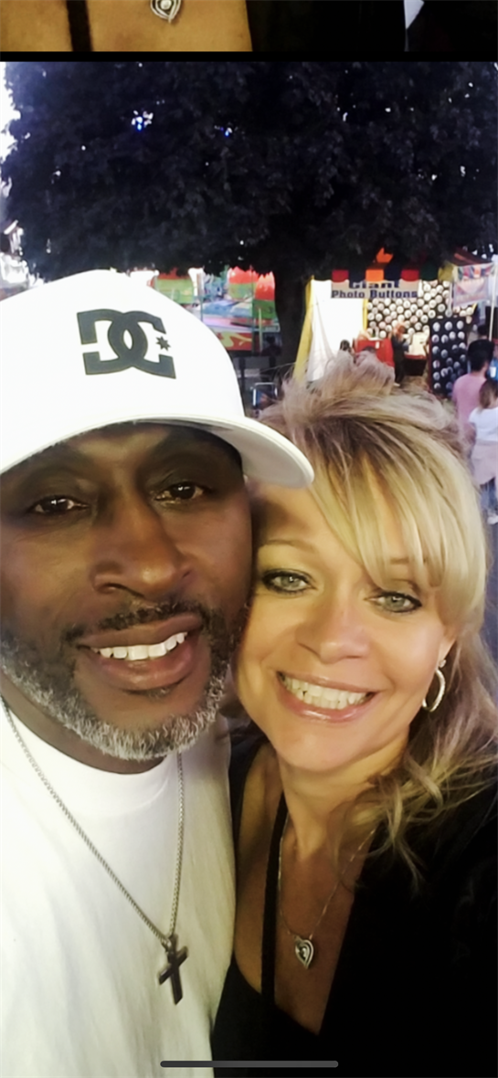 A couple smiles happily together at a summer fair, surrounded by colorful attractions and lights.