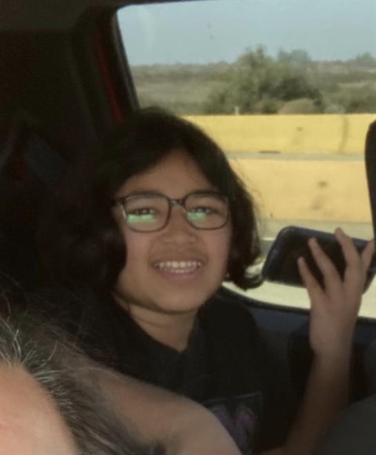 A child with glasses is happily holding a phone while seated in a car, enjoying a road trip.