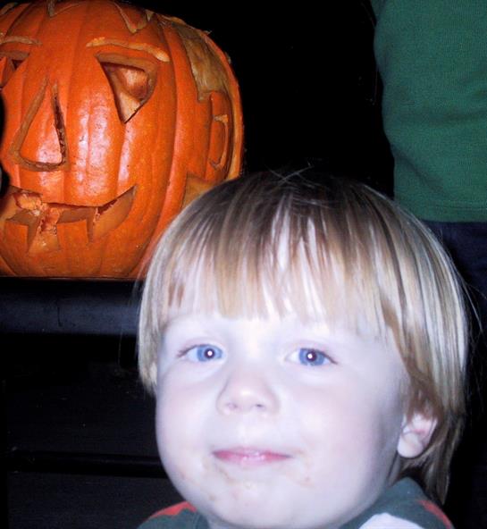 A child with light hair is smiling while posing near a carved pumpkin for Halloween.