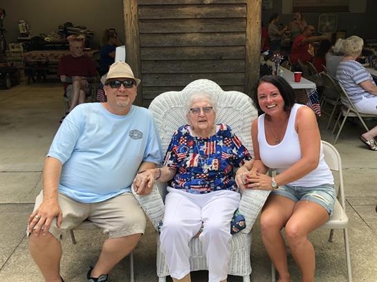 Two adults pose with an elderly woman in a decorative chair at a community event during summer.