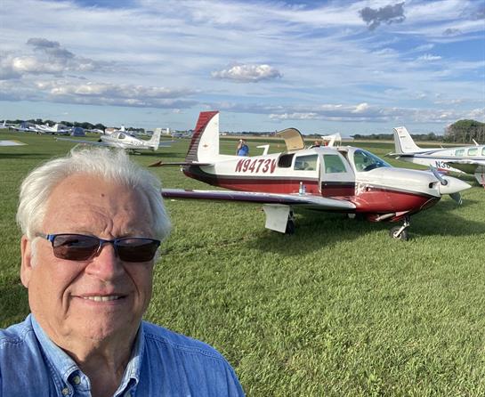 Man enjoys a sunny day at the airfield, posing with a small airplane surrounded by clouds.