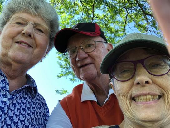 Three seniors smile for a selfie under the bright sunlight, surrounded by greenery in a park.