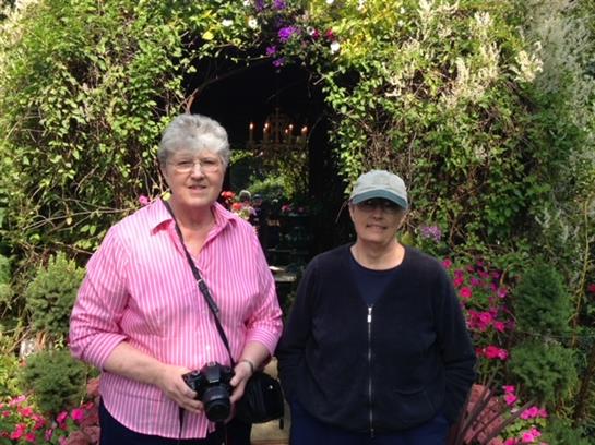 Two women smile while standing amidst colorful flowers and lush foliage in a beautiful garden.