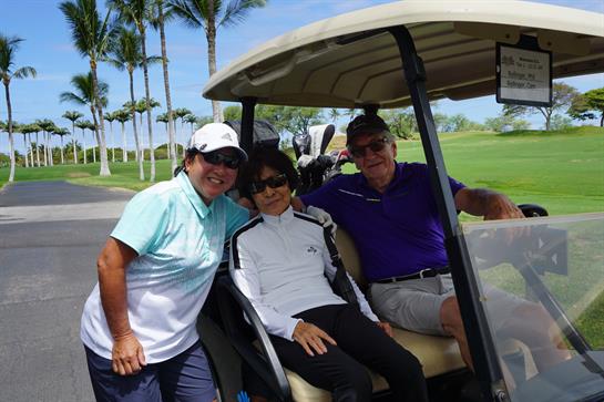 Three people relax in a golf cart, sharing smiles and enjoying the sunny day on the greens.