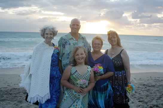 A cheerful family poses together by the ocean as the sun sets, surrounded by colorful flowers.