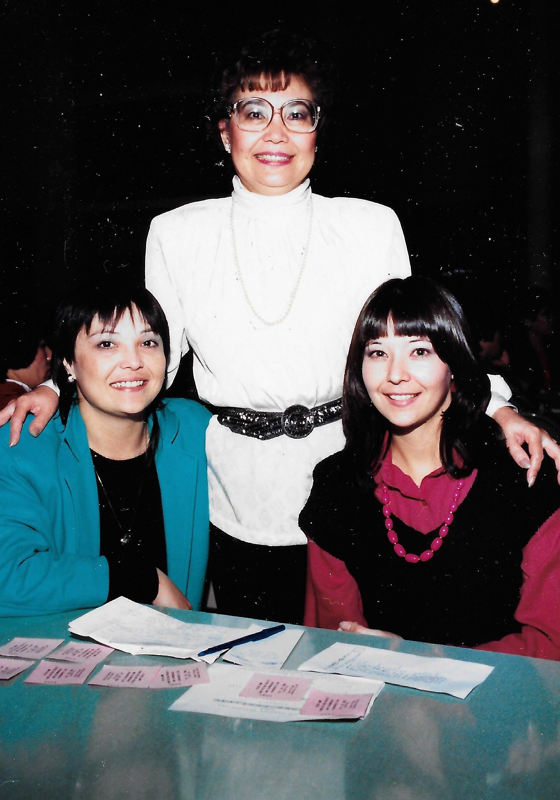 Three women smile as they gather at a social event, dressed elegantly for the occasion.