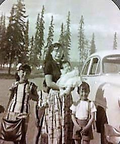 A group of children and an adult woman holding a baby stand by a vintage car in a lush landscape.
